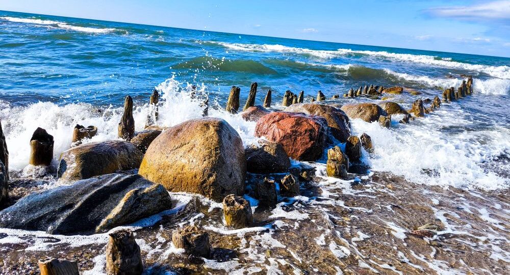 Unter einem strahlend blauen, wolkenverhangenen Ostseehimmel prallen die Wellen gegen große Felsen und alte Holzpfähle am Kühlungsborner Ufer. Die Szene ist lebendig und dynamisch und fängt die Bewegung des Wassers ein - perfekt für eine Auszeit.