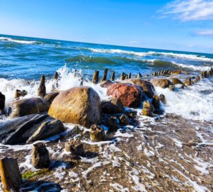 Unter einem strahlend blauen, wolkenverhangenen Ostseehimmel prallen die Wellen gegen große Felsen und alte Holzpfähle am Kühlungsborner Ufer. Die Szene ist lebendig und dynamisch und fängt die Bewegung des Wassers ein - perfekt für eine Auszeit.