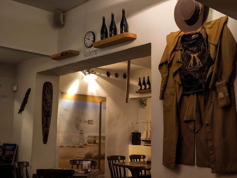 Ein gemütliches Binz-Restaurant mit Holzstühlen, gedämpfter Beleuchtung, Weinflaschen in Regalen, einer Wanduhr und einem Mantel mit Hut an der Wand. Eine Wandmalerei mit einer Outback-Szene ziert die Rückwand.