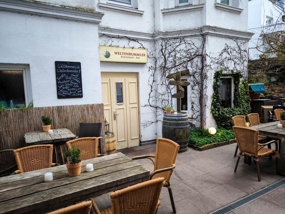 Der Außensitzbereich der Weltenbummler Restaurant Bar in Binz ist mit Holztischen, Korbstühlen, üppigen Topfpflanzen und einer mit Weinreben bedeckten weißen Wand ausgestattet. Eine Speisekarte auf einer Kreidetafel begrüßt die Gäste am Eingang und sorgt für ein charmantes Esserlebnis in Binz.