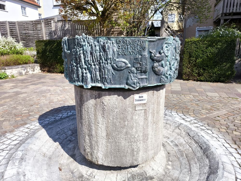 Ein runder Steinbrunnen mit einem bronzenen Reliefband mit Figuren und Gegenständen im gepflasterten Innenhof der Klosterstadt Neresheim. Auf dem Brunnen befindet sich ein Schild mit der Aufschrift Kein Trinkwasser in deutscher Sprache.