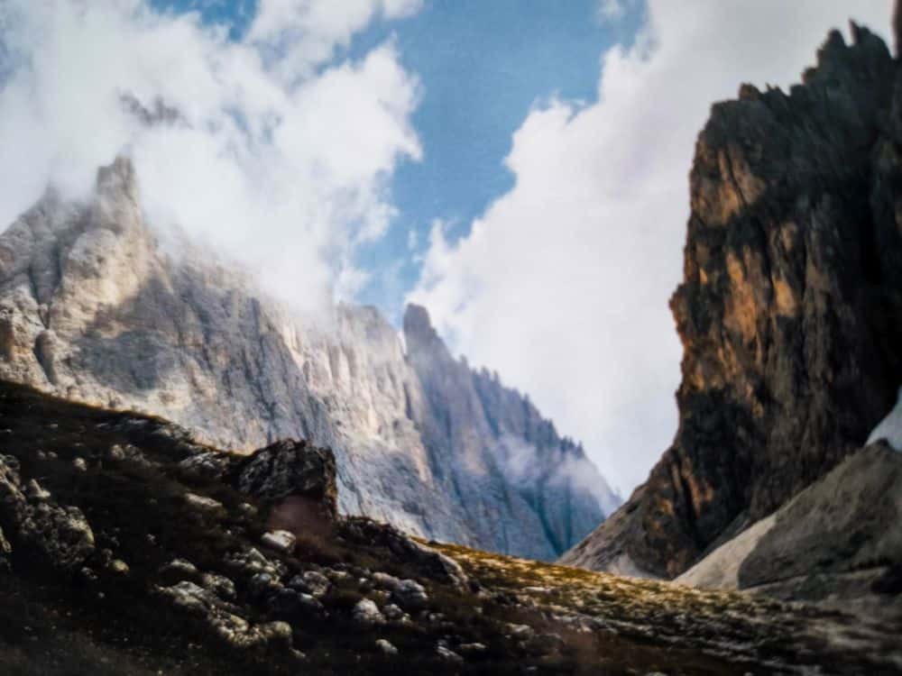 Die felsigen Berggipfel erheben sich steil gegen den teilweise bewölkten blauen Himmel. Das Sonnenlicht beleuchtet die zerklüfteten Felsen der Dolomiten und den schattigen Vordergrund des grasbewachsenen, felsigen Geländes bei der Langkofelscharte.