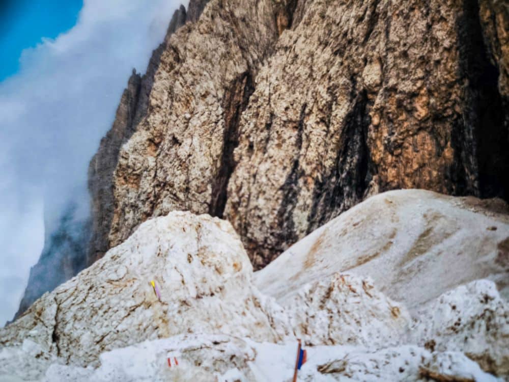 Felsige Berge mit steilen, zerklüfteten Felsen erheben sich über helle, sandige Hänge im Vordergrund. Bunte Wegweiser weisen auf ein klassisches Wandergebiet in den Dolomiten in der Nähe der berühmten Langkofelscharte hin, während Nebelschwaden über die Dolomitenklassiker ziehen.