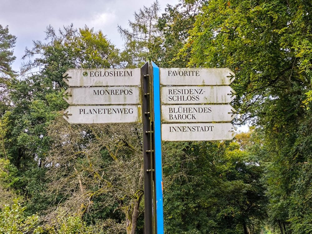Weiße Wegweiser mit schwarzer Schrift weisen auf verschiedene Orte in Ludwigsburg hin, darunter Eglosheim, Monrepos, Planetenweg, Favorite, Residenzschloss, Blühendes Barock und Innenstadt, eingebettet in grüne Bäume.