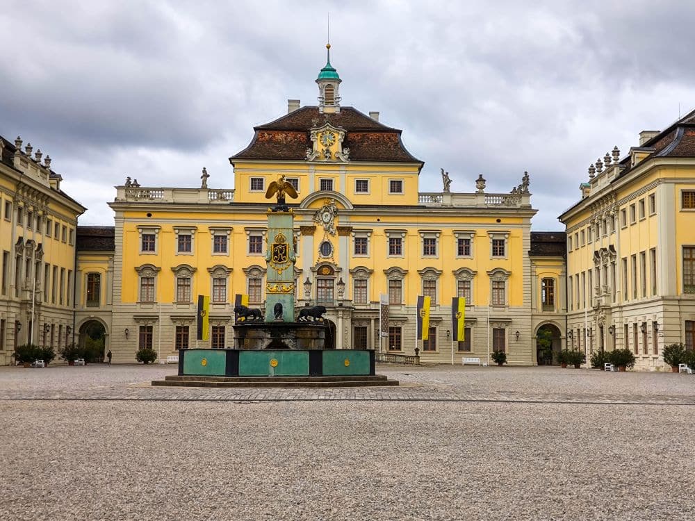 Ein prächtiges, gelbes Barockschloss in Ludwigsburg mit einem zentralen Brunnen in einem weitläufigen Hof mit Kopfsteinpflaster, flankiert von symmetrischen Flügeln, unter einem bewölkten Himmel.