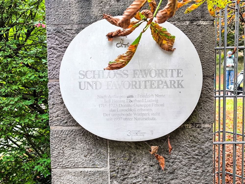 Eine runde weiße Tafel an einer Steinmauer trägt die Aufschrift "Schloss Favorite und Favoritepark" mit zusätzlichem deutschen Text darunter. Gelbbraune Blätter und grünes Laub aus Ludwigsburg rahmen die Tafel teilweise ein.