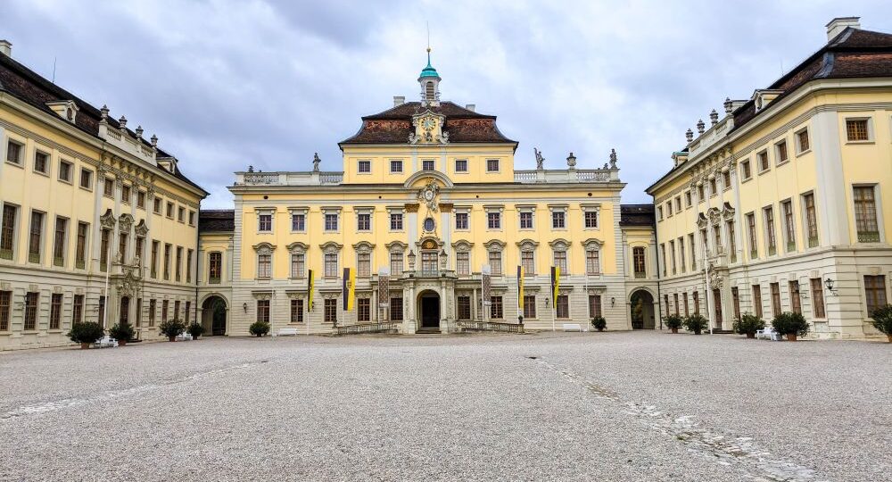 Ein prächtiges gelbes Barockschloss in Ludwigsburg mit zentralem Innenhof, Kiesboden und verzierter Barocke Pracht. Symmetrische Flügel, gewölbte Fenster und bunte Gärten leuchten unter einem wolkenverhangenen Himmel.