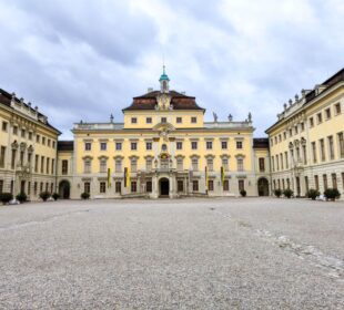 Ein prächtiges gelbes Barockschloss in Ludwigsburg mit zentralem Innenhof, Kiesboden und verzierter Barocke Pracht. Symmetrische Flügel, gewölbte Fenster und bunte Gärten leuchten unter einem wolkenverhangenen Himmel.