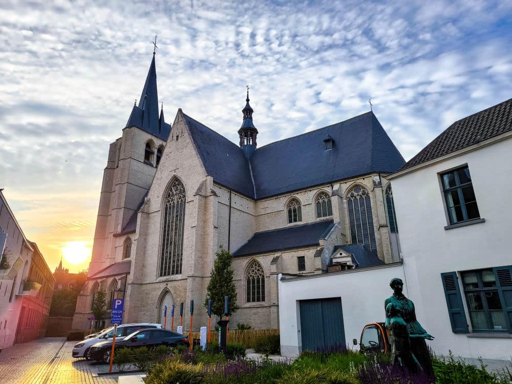 Eine große Steinkirche mit hohen Rundbogenfenstern und einem Glockenturm steht bei Sonnenuntergang unter einem teilweise bewölkten Himmel in der flämischen Stadt. In der Nähe säumen Bronzestatuen, moderne Gebäude und Hotels in Mechelen die Kopfsteinpflasterstraße.