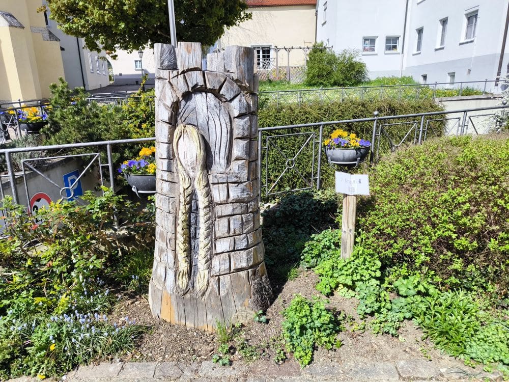 Eine geschnitzte Holzskulptur, die einem Ziegelturm mit einem geflochtenen Zopf in der Mitte ähnelt, steht in einem kleinen Garten in der Klosterstadt Neresheim, umgeben von Grünpflanzen und Blumen. Ein nahe gelegenes Schild und Gebäude im Hintergrund vervollständigen die Szene.