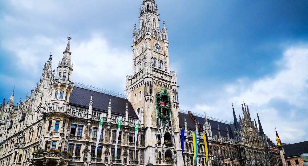 Das Neue Rathaus in München, Deutschland, zeichnet sich durch einen hohen Uhrenturm, eine verschnörkelte, neugotische Architektur und von der Fassade hängende Fahnen vor einem bewölkten Himmel aus.