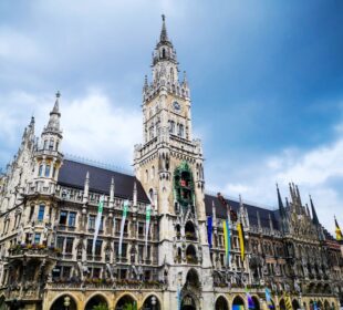 Das Neue Rathaus in München, Deutschland, zeichnet sich durch einen hohen Uhrenturm, eine verschnörkelte, neugotische Architektur und von der Fassade hängende Fahnen vor einem bewölkten Himmel aus.
