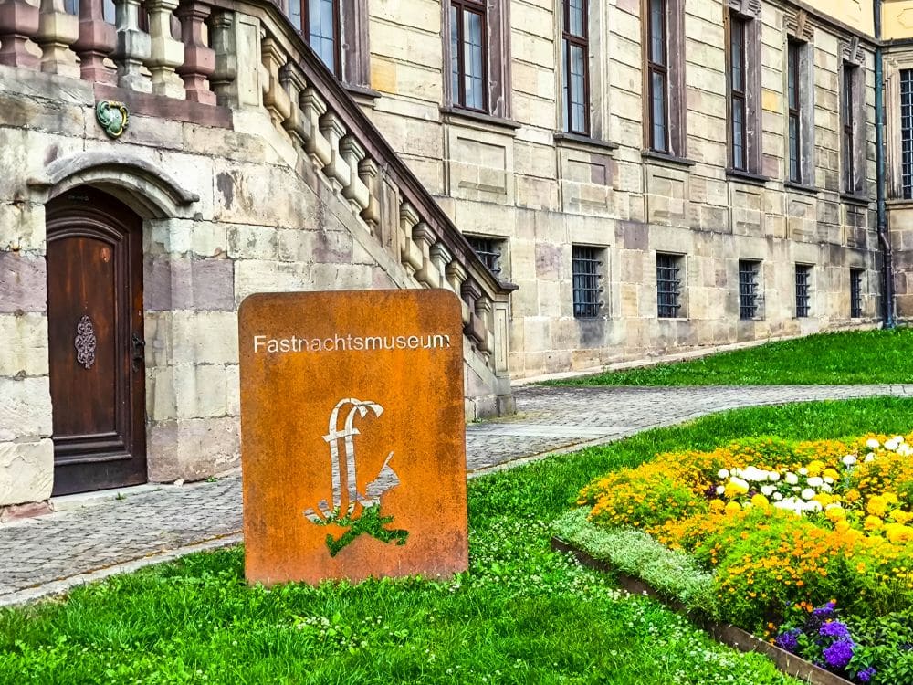 Ein Schild mit der Aufschrift Fastnachtsmuseum steht auf einer Wiese mit bunten Blumen vor einem alten Steingebäude mit gewölbten Fenstern und einer Holztür, das den Charme der historischen Atmosphäre Fuldas einfängt.