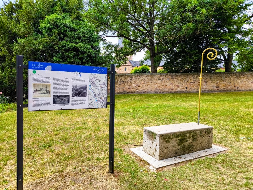 Ein Informationsschild und ein Steinblock mit einem goldenen Stab stehen auf einer Wiese in der Nähe von Bäumen und einer Steinmauer in Fulda, unter einem teilweise bewölkten Himmel.