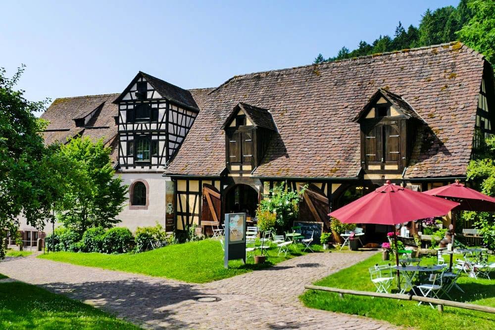 Ein charmantes Fachwerkhaus in Hirsau mit steilem Ziegeldach steht neben einem Straßencafé mit grünen Stühlen und roten Sonnenschirmen, umgeben von üppigem Grün und Bäumen an einem sonnigen Tag.