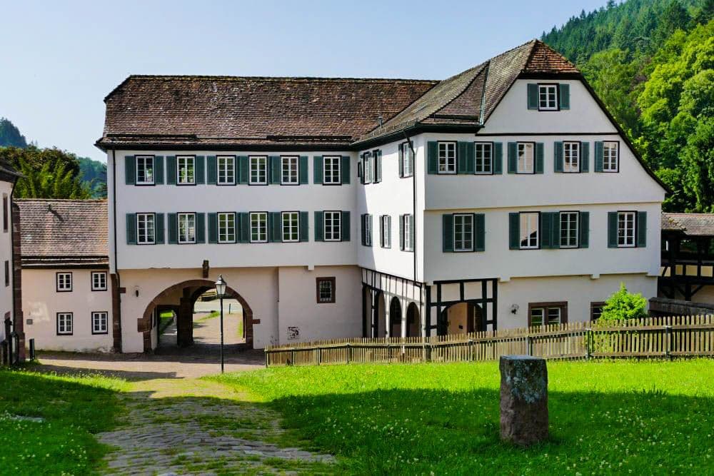 Ein großes, historisches weißes Gebäude mit vielen grün beschlagenen Fenstern, einem bogenförmigen Tor und einem schrägen Ziegeldach steht in Hirsau neben einer Wiese und einem Holzzaun, umgeben von Bäumen und Hügeln.