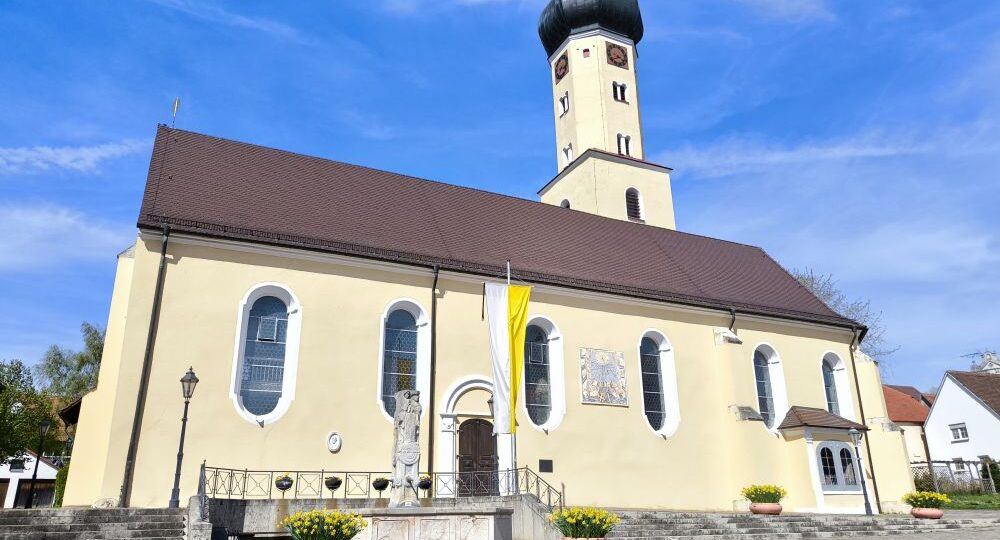 Eine gelbe Kirche mit braunem Dach und einem hohen, achteckigen Uhrenturm steht in der Klosterstadt Neresheim unter blauem Himmel. Stufen führen zum Eingang, der von Blumenkübeln und einer Statue flankiert wird. Neben der Tür hängt eine gelb-weiße Fahne.