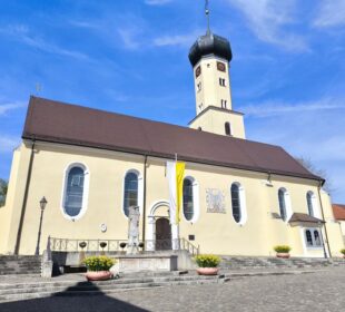 Eine gelbe Kirche mit braunem Dach und einem hohen, achteckigen Uhrenturm steht in der Klosterstadt Neresheim unter blauem Himmel. Stufen führen zum Eingang, der von Blumenkübeln und einer Statue flankiert wird. Neben der Tür hängt eine gelb-weiße Fahne.