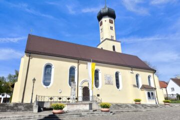 Eine gelbe Kirche mit braunem Dach und einem hohen, achteckigen Uhrenturm steht in der Klosterstadt Neresheim unter blauem Himmel. Stufen führen zum Eingang, der von Blumenkübeln und einer Statue flankiert wird. Neben der Tür hängt eine gelb-weiße Fahne.