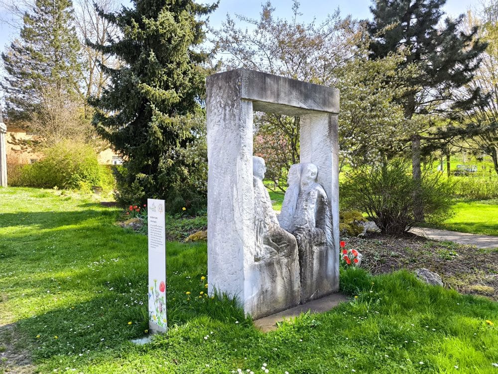 Eine Steinskulptur mit zwei sitzenden menschlichen Figuren in einem rechteckigen Rahmen steht auf einer grünen Wiese in Neresheim, umgeben von Bäumen und Frühlingsblumen. Links in der Umgebung der Klosterstadt ist ein Hinweisschild zu sehen.