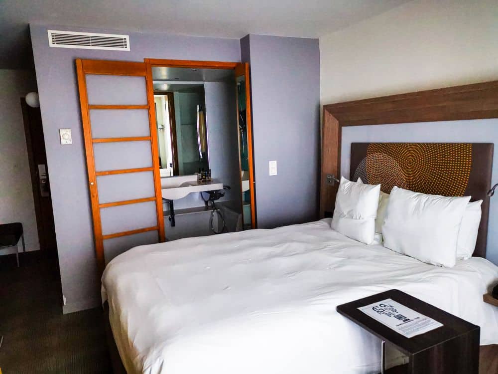 Ein modernes Hotelzimmer in Mechelen mit einem ordentlich gemachten Doppelbett, weißer Bettwäsche, einem hölzernen Kopfteil und einer Schiebetür zum Badezimmer - perfekt für Gäste, die Komfort suchen, während sie diese charmante flämische Stadt erkunden.