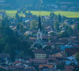 Luftaufnahme von Oberstdorf, einer europäischen Stadt in den Alpen, mit einem hohen Kirchturm in der Mitte, umgeben von Häusern mit roten Dächern, üppigen grünen Bäumen und offenen Feldern im Hintergrund.
