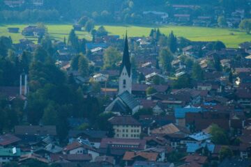 Luftaufnahme von Oberstdorf, einer europäischen Stadt in den Alpen, mit einem hohen Kirchturm in der Mitte, umgeben von Häusern mit roten Dächern, üppigen grünen Bäumen und offenen Feldern im Hintergrund.