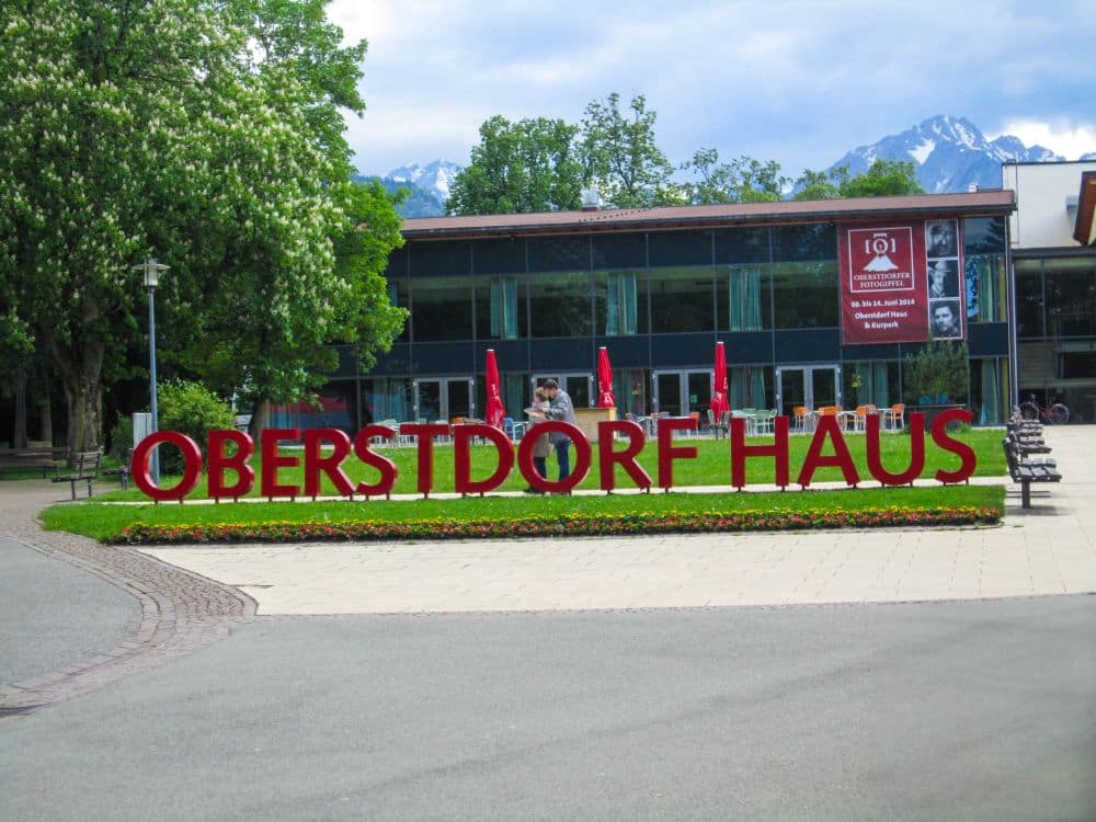 Große rote Buchstaben mit der Aufschrift OBERSTDORF HAUS stehen vor einem modernen Glasgebäude. Im Hintergrund erheben sich Bäume, Bänke und die schneebedeckten Alpenberge von Oberstdorf.