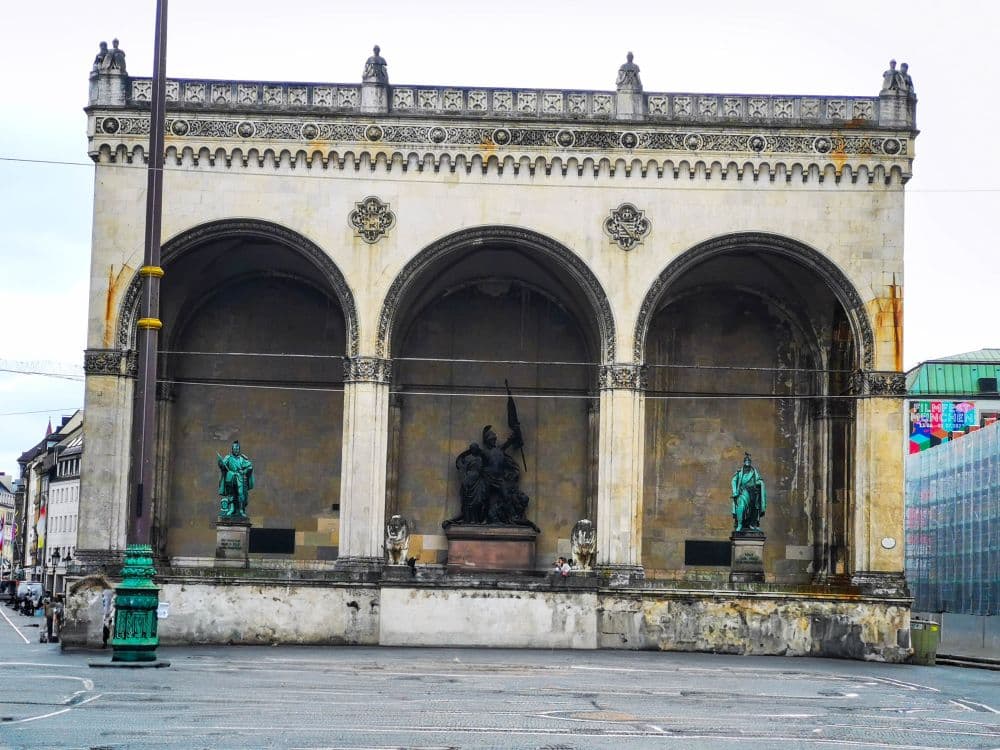 Ein großer historischer Steinbau in München mit drei Bögen, kunstvollen Details entlang des Daches und drei Statuen darunter - eine sitzende Figur in der Mitte, flankiert von zwei stehenden Figuren - auf einem belebten Stadtplatz.