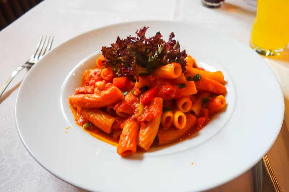 Ein weißer Teller mit Rigatoni, übergossen mit roter Tomatensauce und Blattgemüse, serviert auf einem weißen Tischtuch, dazu eine Gabel und ein Glas gelbes Getränk - klassisches italienisches Flair in einem Erfurter Lieblingsrestaurant.