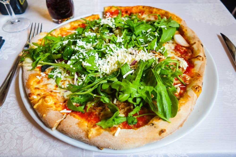 Eine Pizza mit frischem Rucola, geriebenem Käse und Tomatensoße liegt auf einem weißen Teller in einem der Erfurter Lieblingsrestaurants, dazu gibt es Besteck und ein Glas.