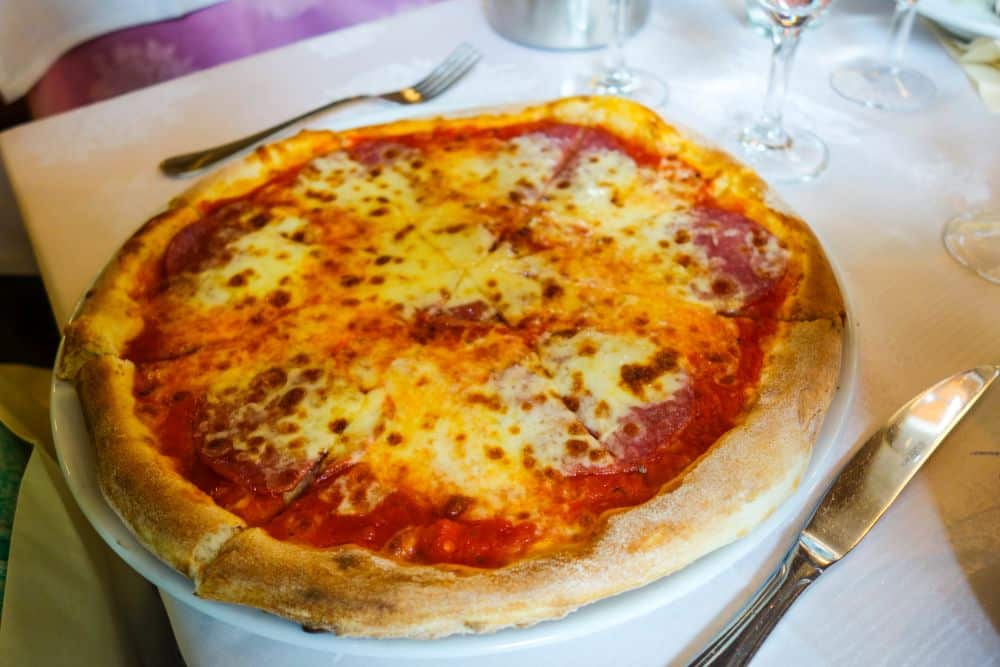 Eine frisch gebackene Käsepizza mit goldener Kruste, Tomatensauce und geschmolzenem Käse auf einem weißen Teller, gedeckt mit Besteck und Weingläsern - wie in einem Erfurter Lieblingsrestaurant.