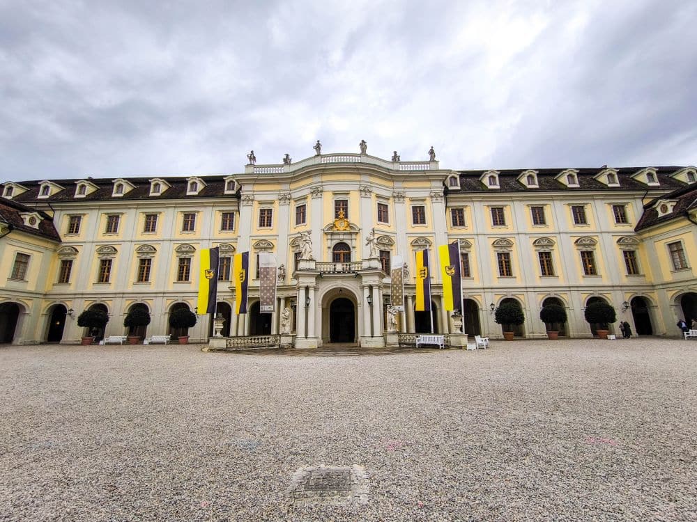 Ein prächtiges, historisches Gebäude mit Bogenfenstern, barocker Pracht und gelb-schwarzen Fahnen steht unter einem wolkenverhangenen Himmel in Ludwigsburg und blickt auf einen weiten Kiesplatz.