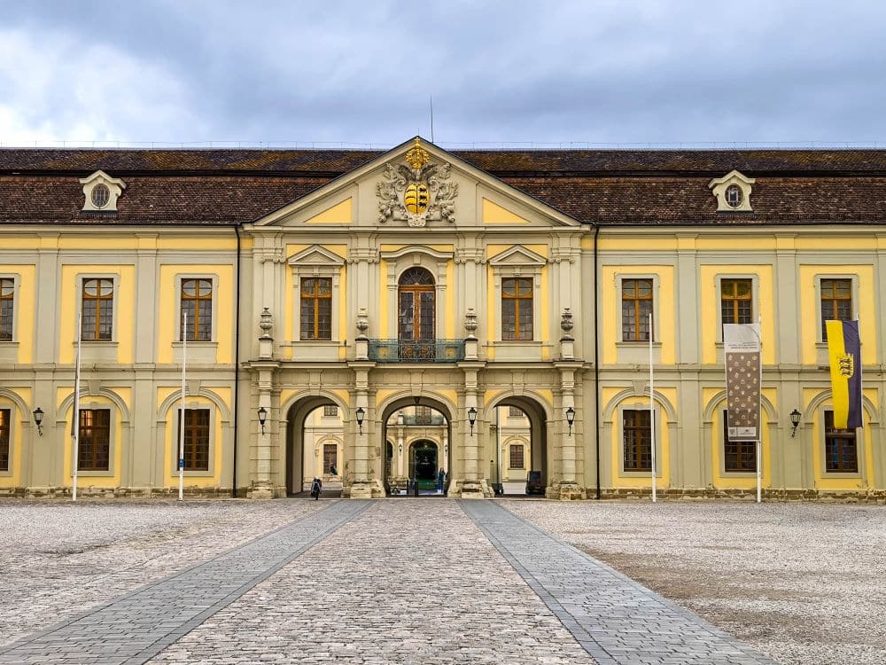 Ein prächtiges gelbes historisches Gebäude in Ludwigsburg mit gewölbten Eingängen, hohen Fenstern und barocker Pracht steht vor einem bewölkten Himmel. Vier Fahnenmasten ragen aus dem steinernen Innenhof davor.