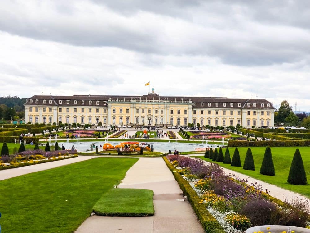 Hinter Bunten Gärten und gestutzten Hecken erhebt sich in Ludwigsburg ein prächtiges Barockschloss mit gelber Fassade, das barocke Pracht ausstrahlt, wenn die Besucher unter wolkenverhangenem Himmel über den Mittelweg flanieren.