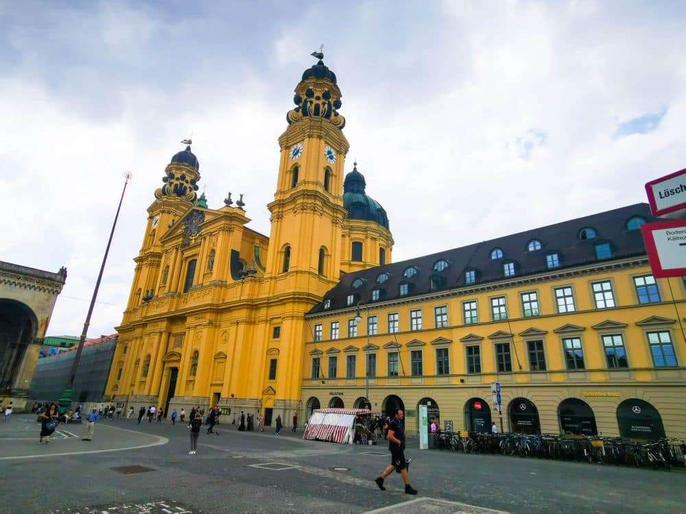 Vor der gelben Fassade der Theatinerkirche in München mit ihren beiden Kuppeltürmen und dem angrenzenden historischen Gebäude laufen und radeln die Menschen bei bewölktem Himmel.