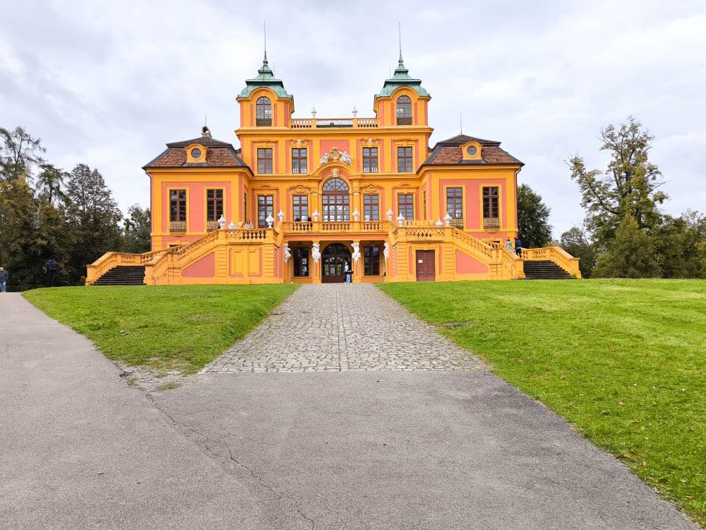 Am Ende eines steinernen Weges, umgeben von Bunten Gärten und Bäumen, steht unter wolkenverhangenem Himmel ein leuchtend gelbes, barockes Herrenhaus mit grünen Kuppeln und kunstvollen Verzierungen, das die Barocke Pracht präsentiert.