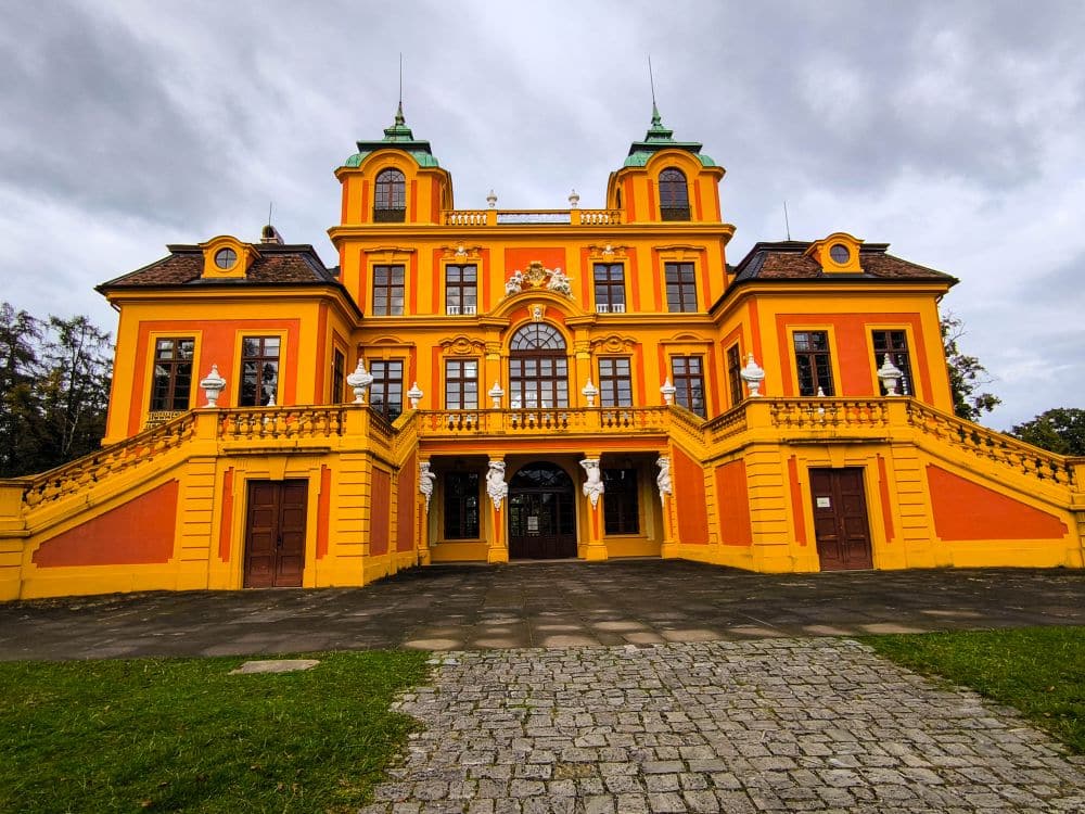 Ein farbenfrohes, in Orange und Gelb gestrichenes Herrenhaus im Barockstil in Ludwigsburg mit symmetrischen großen Treppen, verzierten Fenstern und Türmen mit grünen Dächern, vor einem wolkenverhangenen Himmel, umgeben von Gärten und einem Kopfsteinpflasterweg.
