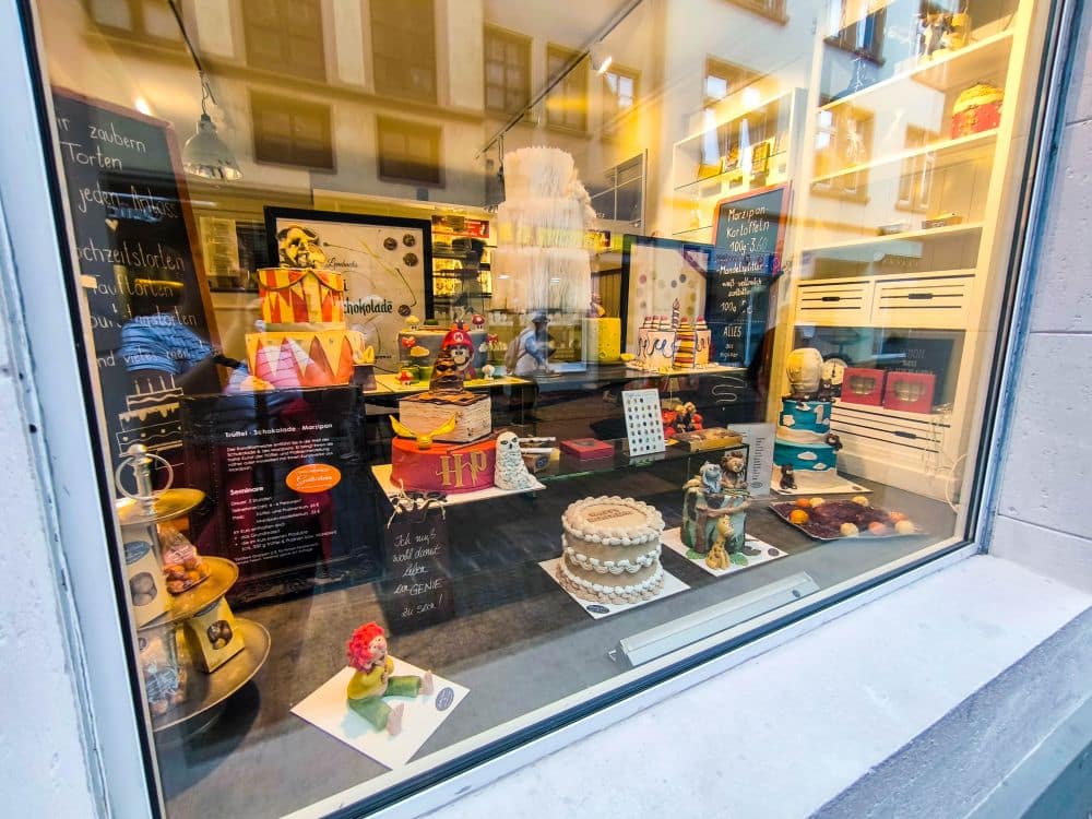 Schaufenster einer Bäckerei in Fulda mit farbenfrohen, aufwändig dekorierten Torten, Gebäck und Dessertständen. Im Glas schimmern Spiegelungen von Menschen und historischen Gebäuden. Der einladende Innenraum wirkt festlich und einladend.