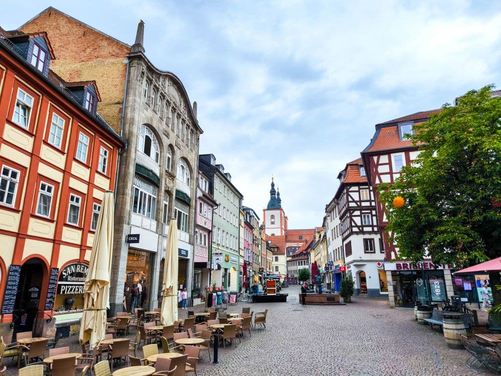 Eine Kopfsteinpflasterstraße in Fulda mit bunten Gebäuden, Straßencafés, Geschäften und einer Kirche mit einem hohen Kirchturm im Hintergrund bei bewölktem Himmel.