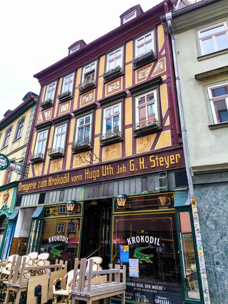 Ein farbenfrohes, historisches, dreistöckiges Gebäude mit kunstvollen Mustern und mehreren Blumenkästen beherbergt im Erdgeschoss ein Geschäft namens Krokodil in Steyr, Österreich - eine Reminiszenz an die charmanten Straßen in Fulda. Am Eingang stehen Stühle für den Außenbereich bereit.