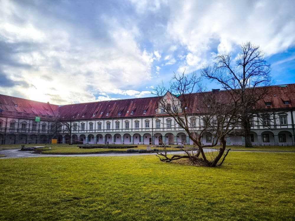 Ein historisches, weißes Barockgebäude mit roten Dachziegeln, Teil des Klosters Benediktbeuern, umgibt einen grasbewachsenen Innenhof mit einem blattlosen Baum unter einem teilweise bewölkten Himmel und mit Blick auf die nahe gelegenen Berge. Die Szene ist friedlich und von sanftem, natürlichem Licht durchflutet.