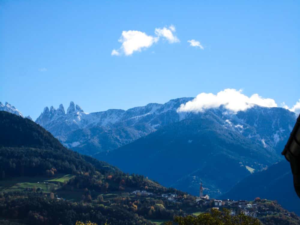 Eine malerische Berglandschaft in Südtirol mit schneebedeckten Gipfeln, bewaldeten Hängen und dem Dorf Feldthurns, das sich im Tal unter einem strahlend blauen Himmel befindet - perfekt, um den berühmten Keschtnweg zu erkunden.