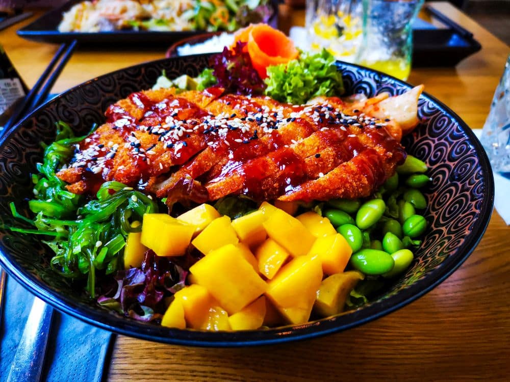Eine farbenfrohe Schale mit gegrilltem Hähnchenfleisch mit Sesam, gewürfelter Mango, Edamame, Seetang-Salat, Blattgemüse und Karotten, serviert auf einem Holztisch, aus einem der Erfurter Lieblingsrestaurants.