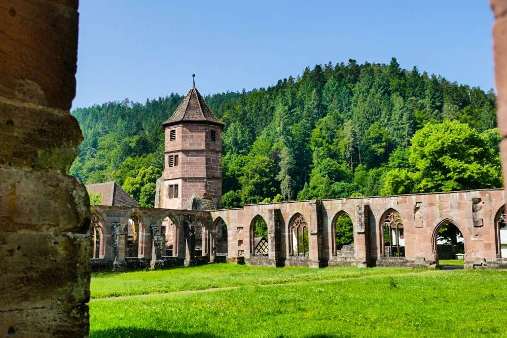 Ruinen der Hirsauer Backsteinabtei mit Rundbogenfenstern und einem zentralen Turm, umgeben von grünem Gras und dicht bewaldeten Hügeln unter einem klaren blauen Himmel.