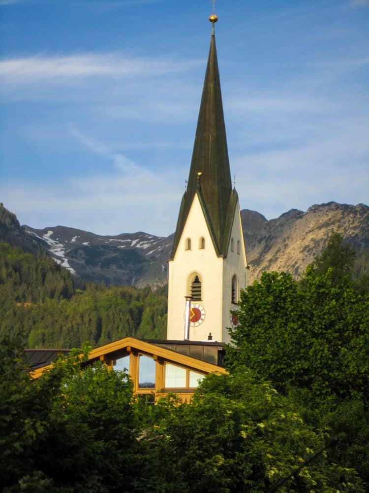 Eine hohe Kirche mit spitzem Kirchturm erhebt sich über einem von grünen Bäumen umgebenen Holzhaus, das sich unter blauem Himmel in die bewaldeten Hügel und schroffen Alpenberge bei Oberstdorf einfügt.