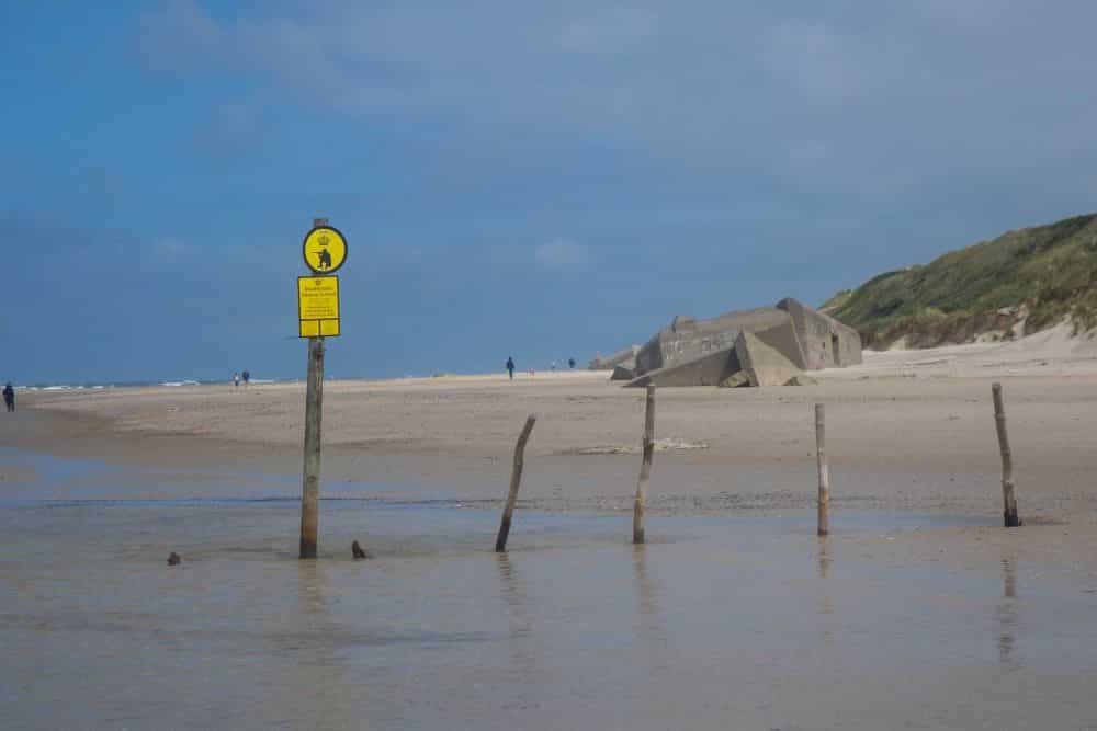 An einem Sandstrand in Westdänemark stehen ein Warnschild an einem Pfahl und Holzpfähle im flachen Wasser. Im Hintergrund spazieren Menschen in der Nähe einer Betonbunkerruine und grasbewachsenen Sanddünen unter dem teilweise bewölkten Himmel von Blavand.