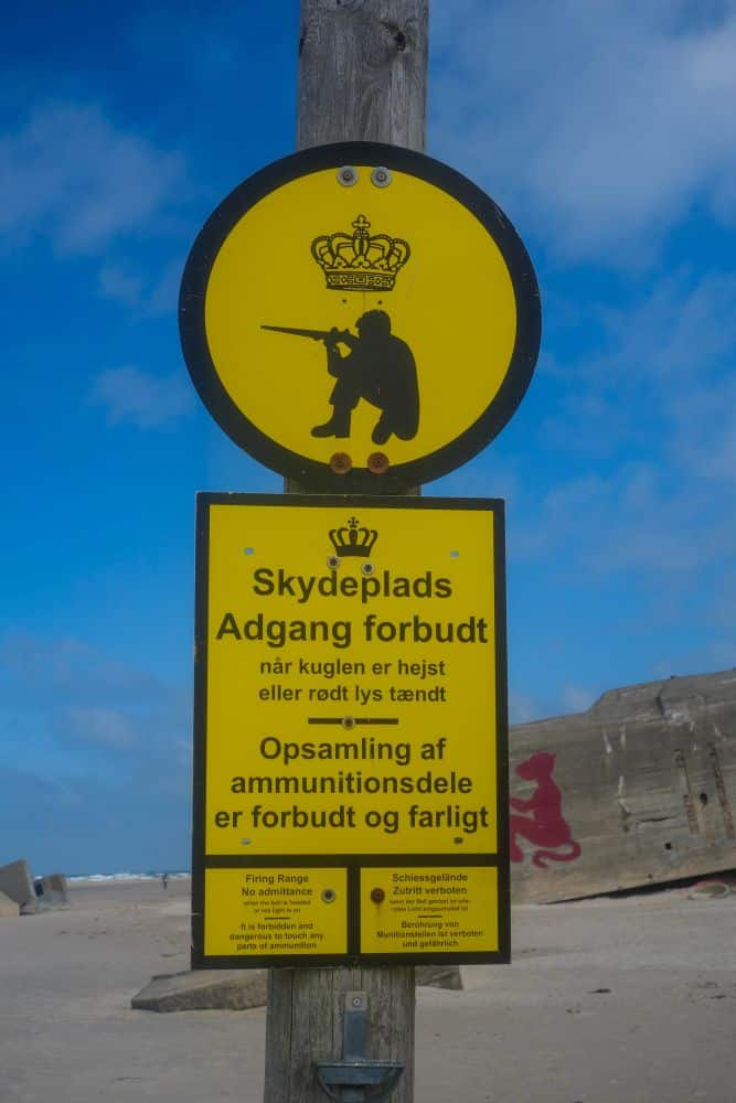 Ein gelbes Warnschild an einem Strand in Blavand, Dänemarks Westen, zeigt die Silhouette einer Person, die mit einem Gewehr zielt, mit dänischem Text, der vor Zugangsbeschränkungen und Gefahren warnt; im Hintergrund ist ein großer Bunker zu sehen.