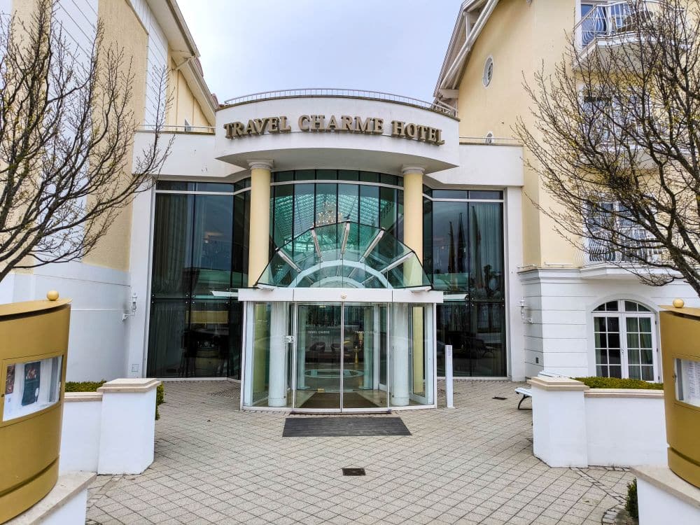Der Eingang des Travel Charme Hotels in Binz besteht aus Glastüren, einer geschwungenen Glasmarkise und großen Fenstern, die von hellen Wänden und kahlen Bäumen auf beiden Seiten eingerahmt werden.