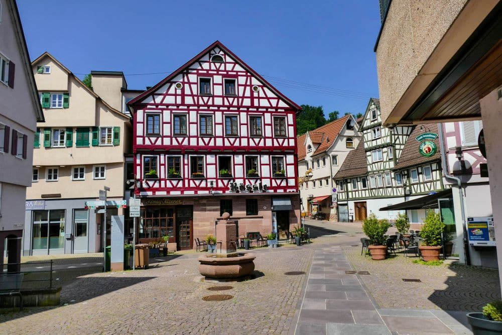 Eine malerische Calwer Straße mit Fachwerkhäusern, einem kopfsteingepflasterten Platz, Topfpflanzen und einem runden Steinbrunnen in der Mitte, und das alles unter einem klaren blauen Himmel.